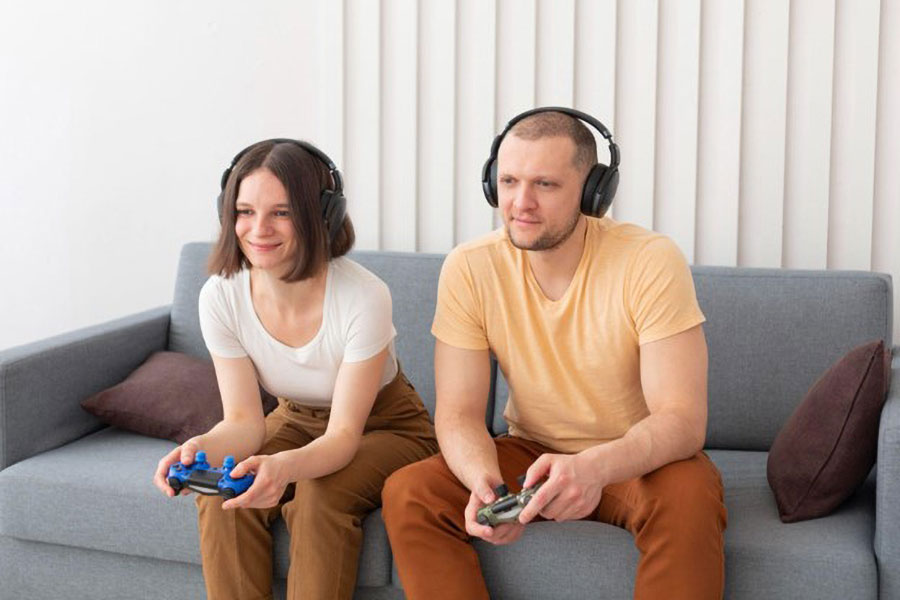 Two gamers sitting on a couch playing games with their controllers and their headphones.